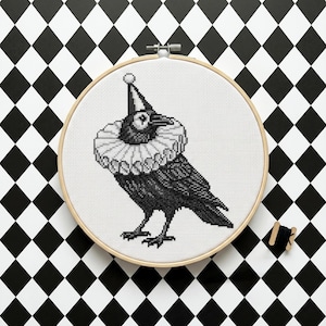 May include: A black and white cross-stitch of a crow wearing a ruffled collar and a pointed hat, set in a wooden embroidery hoop. The crow is on a white background, with a black and white diamond pattern in the background. A spool of black thread is next to the hoop.