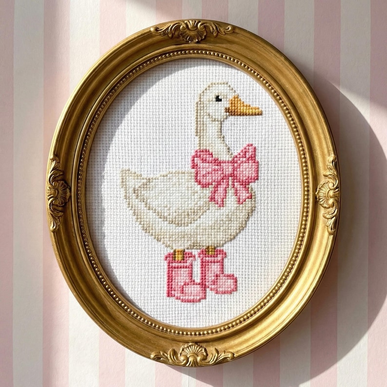 May include: A framed cross-stitch artwork featuring a white goose with a pink bow and boots. The goose has a yellow beak and is set against a white background. The frame is gold-colored and oval-shaped.