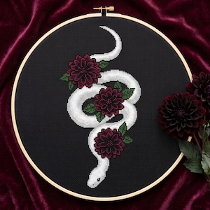 May include: A cross-stitch embroidery piece in a round wooden hoop featuring a white snake winding through three dark red flowers with green leaves, all stitched onto a black fabric background, creating a high-contrast decorative wall art item.