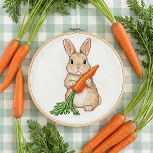 Könnte beinhalten: Ein Kreuzstich-Stickrahmen mit einem braunen Hasen, der eine orangefarbene Karotte mit grünen Blättern hält. Frische Karotten mit grünem Kraut umgeben den Rahmen, auf einer grün-weiß karierten Oberfläche.