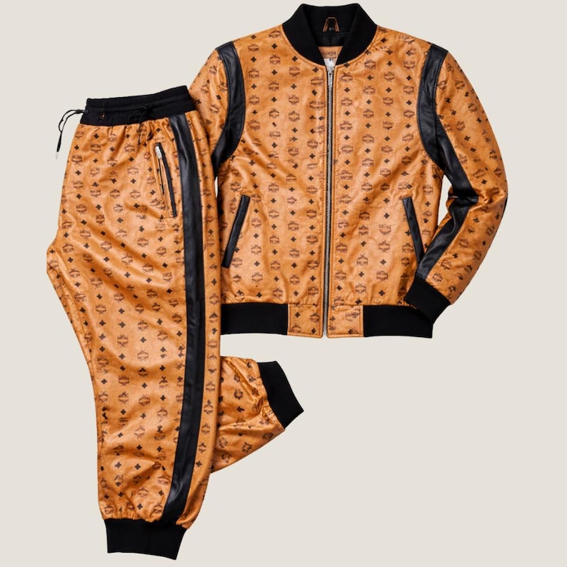 K&ouml;nnte beinhalten: Ein zweiteiliges Trainingsanzug-Set in einem beigefarbenen Farbton mit sich wiederholendem Muster. Die Jacke hat schwarze Akzente an den &Auml;rmeln und am Kragen sowie einen Rei&szlig;verschluss. Die Hose hat einen schwarzen Streifen an der Seite und einen Kordelzug in der Taille.