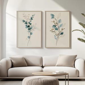 Könnte beinhalten: Zwei vertikale gerahmte Wandbilder mit botanischen Aquarellillustrationen von Eukalyptuszweigen in Blaugrün-, Blau- und Brauntönen auf cremefarbenem Hintergrund, die nebeneinander in hellen Holzrahmen an einer Wand hängen.