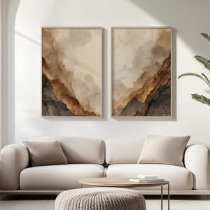 May include: Two rectangular wall art prints in light-colored frames featuring abstract watercolor-style layers of cream, tan, brown, and dark charcoal gray. The artwork displays flowing, organic shapes that resemble layered geological formations.