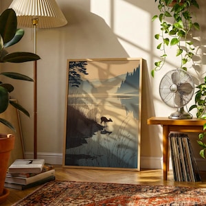 May include: A framed landscape art print depicting a deer by a lake with mountains in the background. The artwork is in a wooden frame and is displayed in a room with a floor lamp, plants, and a vintage fan. The color palette includes shades of blue, brown, and beige.