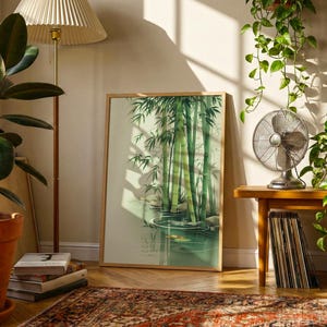 Op de afbeelding: Een ingelijste kunstdruk met een aquarel van bamboestengels in een serene vijver. Het kunstwerk wordt getoond in een kamer met een staande lamp, potplanten en een vintage ventilator op een houten tafel. De kamer heeft een warme, uitnodigende sfeer.