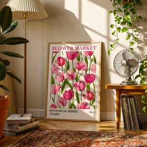 May include: A framed art print featuring a floral design of pink tulips. The print is labeled "FLOWER MARKET" and "BOTANICAL SERIES TULIPS." The artwork is displayed in a room with a lamp, plants, and a small table with a fan.