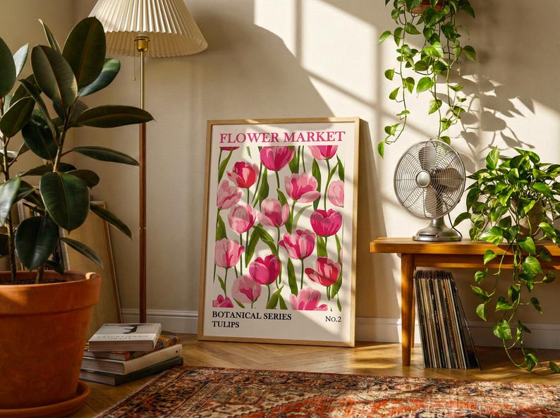 May include: A framed art print featuring a floral design of pink tulips. The print is labeled "FLOWER MARKET" and "BOTANICAL SERIES TULIPS." The artwork is displayed in a room with a lamp, plants, and a small table with a fan.