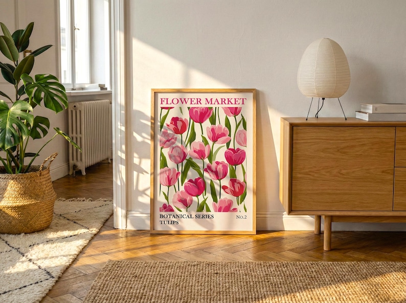 May include: A framed print with a floral design of pink tulips. The print is labelled "Flower Market" and "Botanical Series Tulips." The artwork is displayed in a room with a wooden cabinet, a woven basket, and a decorative lamp.