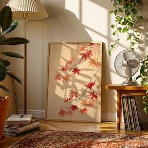May include: A framed art print depicting red and orange maple leaves on branches. The artwork is in a room with a floor lamp, potted plants, and a vintage fan on a wooden table. The room has a warm atmosphere, with a patterned rug and sunlight streaming in.