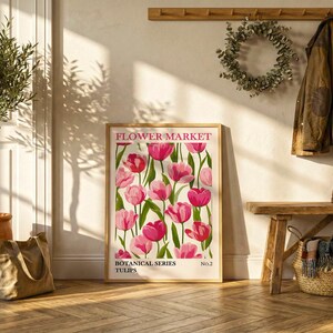 May include: A framed "Flower Market" poster featuring pink tulips with green stems and leaves. The poster is part of the "Botanical Series" and is displayed near a potted olive tree, a tote bag, and a wooden bench with a coat and boots.