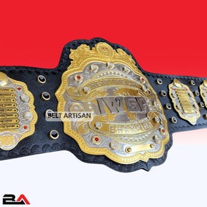 Può includere: Una cintura da campionato oro e argento con la scritta "HEAVYWEIGHT CHAMPION" e "IWGP" sul davanti. La cintura ha un cinturino in pelle nera con dettagli dorati e gioielli rossi. Sono visibili anche le parole "BELT ARTISAN".