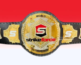 Op de afbeelding: Een kampioenschapsriem met een zwarte leren band en een grote, ronde, goudkleurige middenplaat. De plaat toont de tekst "WORLD CHAMPION" en een centraal schild met een wit "S"-logo en de rode tekst "strikeforce" eronder.