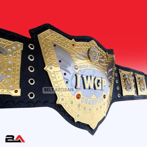 Può includere: Una cintura da campionato di wrestling con una grande placca centrale dorata e argentata con la scritta IWGP WORLD HEAVYWEIGHT CHAMPION. La cintura presenta un cinturino in pelle nera con placche laterali dorate, rivetti decorativi e piccole gemme rosse.