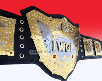 Pode incluir: Um cinturão de campeonato de luta livre com uma grande placa central dourada e prateada com o texto IWGP WORLD HEAVYWEIGHT CHAMPION. O cinto tem uma tira de couro preta com placas laterais douradas, rebites decorativos e pequenas gemas vermelhas.
