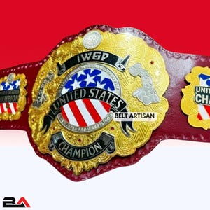 Può includere: Una cintura da campionato di wrestling dorata e rossa con la scritta "UNITED STATES CHAMPION" e "IWGP". La cintura presenta un emblema centrale con un disegno a stelle blu e bianche e le parole "BELT ARTISAN".