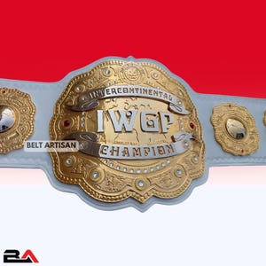 May include: Gold and silver Intercontinental Championship belt with the text "INTERCONTINENTAL IWGP CHAMPION" on a white leather strap. The belt has intricate detailing and is set against a red and white background.