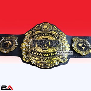Può includere: Una cintura da campionato nera e dorata con la scritta "Heavyweight IWGP Champion" e "Belt Artisan". La cintura presenta intricati dettagli dorati, tra cui un'emblema del globo e il nome "Kazuchika Okada".