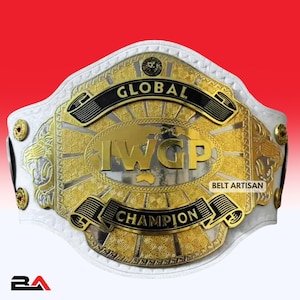 Può includere: Una cintura da campionato in pelle bianca con una grande placca centrale dorata e argentata con la scritta GLOBAL IWGP CHAMPION. La placca presenta intricati motivi incisi, teste di leone laterali e piccoli borchie circolari dorate.