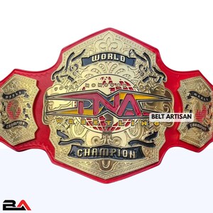 Puede incluir: Un cinturón de campeonato de lucha libre dorado y rojo con el texto "TNA Wrestling Champion" y "World Champion". El cinturón tiene detalles intrincados y está hecho de metal. Las palabras "Belt Artisan" también son visibles.