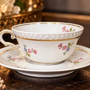 Könnte beinhalten: Ein weißes Teetassen- und Untertassenset aus Porzellan mit einem zarten Blumenmuster aus kleinen rosa und blauen Blüten. Der Rand der Tasse und der Untertasse ist mit Gold verziert, und beide Teile weisen ein strukturiertes, geprägtes Randdetail auf.