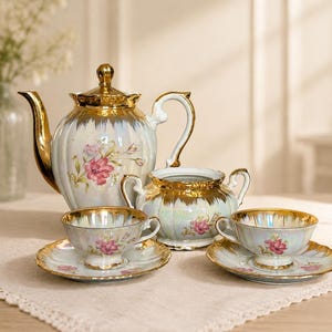 Peut inclure: Un service à thé en porcelaine comprenant une théière, un sucrier et deux tasses avec leurs soucoupes assorties. Les pièces présentent une finition irisée, des bords et anses dorés, et un motif floral rose sur fond blanc.