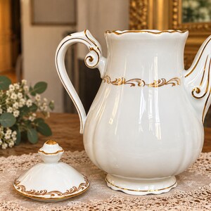 Może przedstawiać: Biały porcelanowy imbryk z żebrowanym korpusem i pasującą pokrywką. Imbryk posiada złote wykończenia wzdłuż falistego brzegu, uchwytu, dzióbka i podstawy, z delikatnym złotym wzorem filigranowym otaczającym górną część korpusu naczynia.