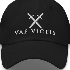 May include: A black baseball cap featuring white embroidery on the front. The design consists of two crossed swords positioned above the text VAE VICTIS, which is stitched in a clean, capitalized, serif font centered on the crown of the hat.