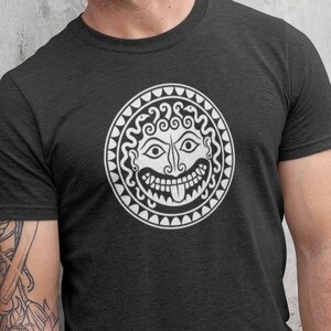 Puede incluir: Una camiseta de manga corta en color gris carbón oscuro con un gráfico circular blanco en el pecho. El diseño muestra una cabeza de Medusa estilizada con serpientes por cabello, ojos grandes y la lengua fuera, dentro de un borde decorativo.