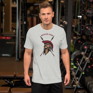 Puede incluir: Camiseta gris claro con un gráfico de un casco espartano, lanzas cruzadas y el texto "MOLON LABE" en borgoña. El casco es de bronce con una cresta roja. La camiseta de manga corta está hecha de un material suave.