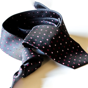 May include: A black tie with a repeating pattern of small, pink squares. The tie is made of a textured fabric and is displayed on a white surface. The tie is rolled up, showing the front and back of the fabric.