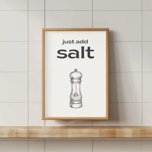 May include: A framed print with the words "just add salt" above a sketch of a salt grinder. The artwork is in a wooden frame and is displayed on a wooden shelf against a white tiled wall.