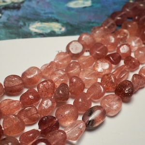 May include: Strands of polished, reddish-orange strawberry quartz beads. The beads are irregularly shaped and translucent, with a speckled appearance. The background features a blurred image of a water lily painting. These beads are suitable for jewelry making.