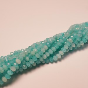 May include: Strands of faceted, light blue beads. The beads are small and round, with multiple facets that catch the light. The beads are strung together, forming a long, flowing strand. The background is a soft, neutral color.
