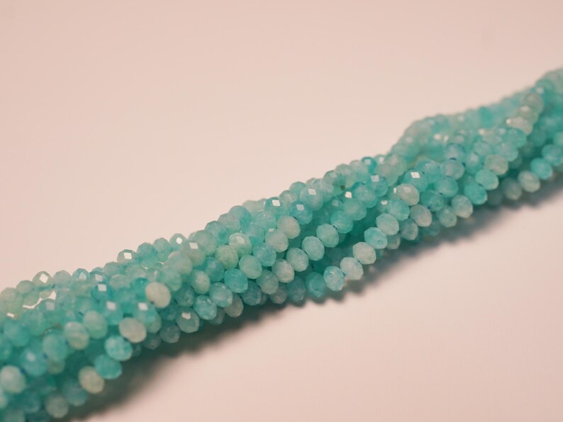 May include: Strands of faceted, light blue beads. The beads are small and round, with multiple facets that catch the light. The beads are strung together, forming a long, flowing strand. The background is a soft, neutral color.