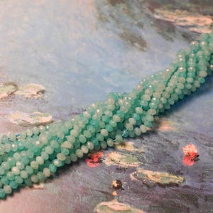 May include: Strands of faceted, light blue beads. The beads are small and round, with a sparkling, polished finish. The beads are strung together, forming a long, flowing strand. The background is a painting with blue and green tones.