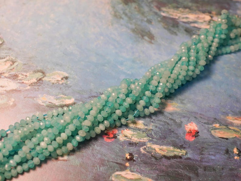 May include: Strands of faceted, light blue beads. The beads are small and round, with a sparkling, polished finish. The beads are strung together, forming a long, flowing strand. The background is a painting with blue and green tones.