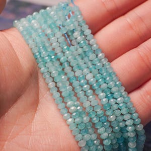 May include: Strands of small, faceted, light blue gemstone beads. The beads are strung together, creating a delicate and shimmering effect. The beads are held in a hand, showcasing their color and texture. Ideal for jewelry making.