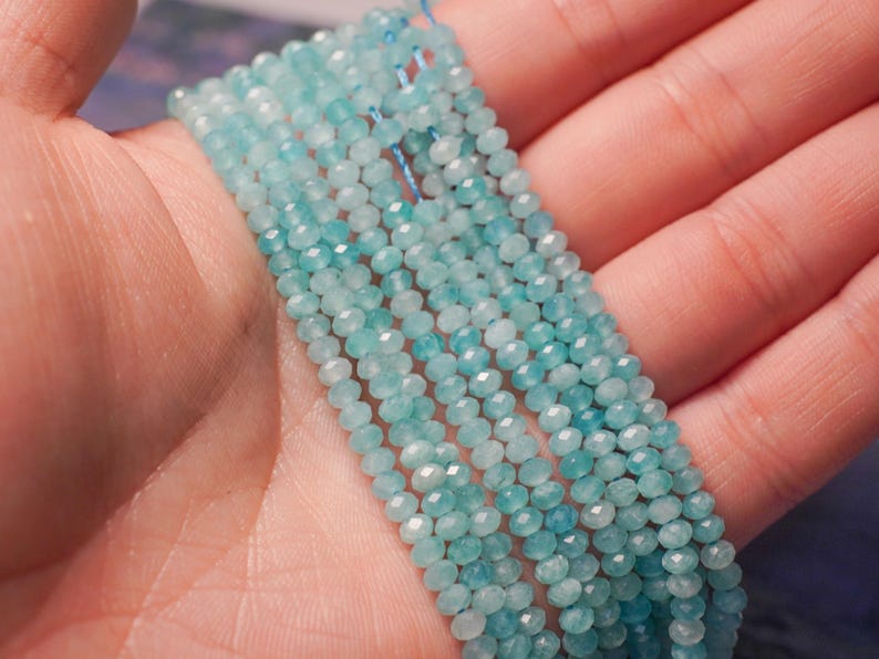 May include: Strands of small, faceted, light blue gemstone beads. The beads are strung together, creating a delicate and shimmering effect. The beads are held in a hand, showcasing their color and texture. Ideal for jewelry making.