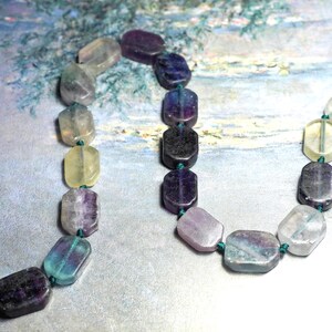 May include: A necklace of rectangular fluorite beads in various colours, including purple, blue, yellow, and clear. The beads are strung on a green cord. The necklace is approximately 40 cm long.