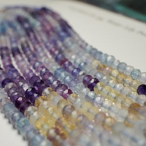 May include: Strands of faceted gemstone beads in shades of purple, blue, and yellow. The beads are strung together, creating a flowing, textured appearance. The gemstones are likely used for jewelry making.