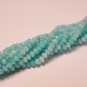 May include: Strand of faceted, round beads in shades of light blue and white. The beads are strung together, creating a textured, flowing appearance. The beads are likely made of a gemstone or glass, perfect for jewelry making.