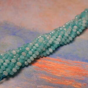 May include: Strand of faceted, light blue beads. The beads are small and round, with a slightly iridescent quality. The beads are strung together, forming a long, flexible strand. The background is a mix of blue, orange, and pink.