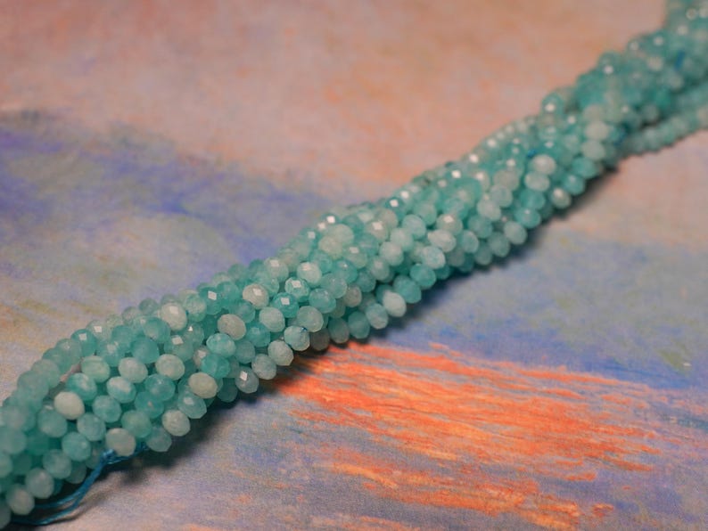 May include: Strand of faceted, light blue beads. The beads are small and round, with a slightly iridescent quality. The beads are strung together, forming a long, flexible strand. The background is a mix of blue, orange, and pink.