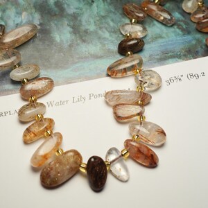 May include: Close-up of a necklace featuring oval-shaped, translucent beads with reddish-brown inclusions. The beads are strung together with gold-colored connectors. The necklace is approximately 36.375 inches (92.4 cm) long. The text "Water Lily Pond" is visible.