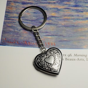 May include: Silver heart-shaped locket keyring with detailed floral patterns. The locket features a small heart in the centre and is suspended from a chain and a ring. The background includes text and a painting.