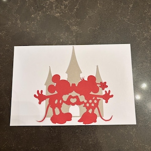 May include: A red paper cutout of Mickey and Minnie Mouse kissing, with a heart between them. The silhouettes are in front of a tan castle on a white background. The image is on a dark brown surface.