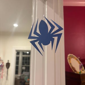 Op de afbeelding: Een blauwe spinnensticker is bevestigd aan een witte kolom. Het spinnendesign heeft een centraal lichaam en acht puntige poten. De achtergrond toont een kamer met een deur en een decoratief item.