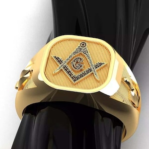May include: A gold-colored ring with a rectangular face featuring a Masonic symbol. The ring has a polished finish and is displayed on a black surface. The Masonic symbol includes a compass, square, and the letter "G".