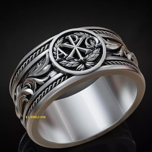 Könnte beinhalten: Ein silberfarbener Ring mit aufwendigem Design. Der Ring hat ein zentrales Emblem in einem kreisförmigen Rahmen, umgeben von detaillierten floralen und seilartigen Mustern. Der Ring ist breit und poliert und reflektiert das Licht.