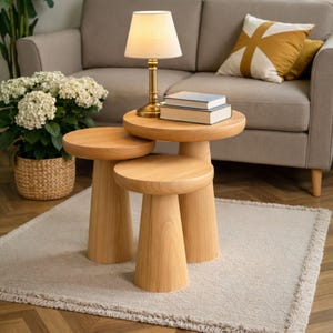 May include: Three wooden side tables with a natural finish. A small lamp with a beige shade and books are on the largest table. A beige sofa and a potted plant are in the background. The tables are approximately 40 cm in diameter.
